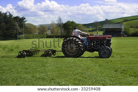 a man cutting a rugby fields grass with a tractor and gang mower looking behind, showing the grass cuttings in the air behind the mower.