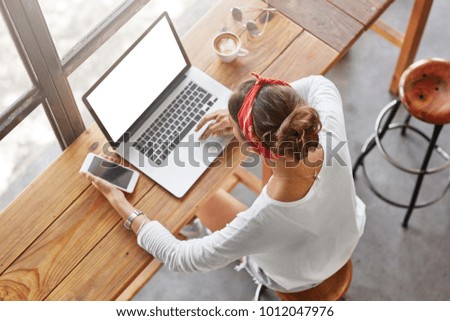 Top view of fashionable femle student learns online on laptop computer, prepares for classes or exam, holds cell phone as waits for call, spends free time at cafeteria with mug of tasty latte