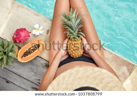 Top view of slim woman with tanned skin, sits near hotel pool enjoy vegan healthy diet food and eat tropical fruits, has summer pool party. Female eats juicy exotic yield: pineapple and papya