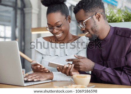 Black woman and man have informal meeting, glad to view photos on smart phone, wear spectacles, work together at common project via laptop computer, drink coffee. People and technology concept