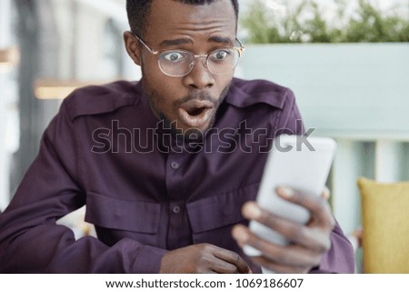 Shocked dark skinned young man in spectacles surprised as gets bill receipt on mobile phone, stressed to read shocking news, has business problems, reacts emotionally, worried about something