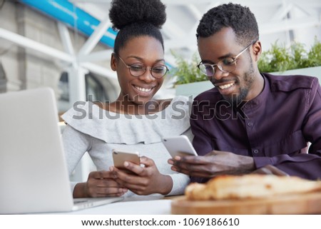 Smiling African American friends meet together at cafe, use modern technologies for entertainment. Dark skinned delighted young female and male hold smart phones, download new applications or files
