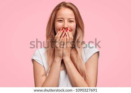 Horizontal shot of good looking young female giggles joyfully, covers mouth as tries stop laughing, wears casual white t shirt isolated on pink background. Happy woman recieves proposal from boyfriend