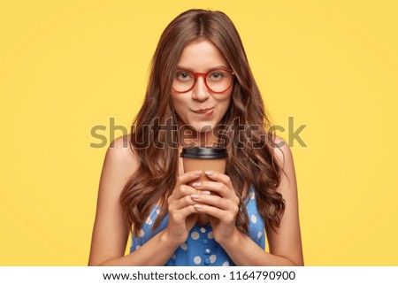 Indoor shot of pretty girl with intriguing expression, purses lower lip, holds paper cup of coffee, waits for suggestion from guy to date, wears round spectacles, isolated over yellow background