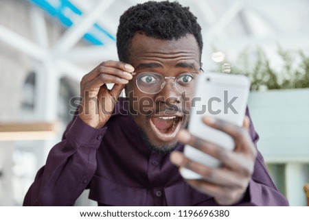 Shocked dark skinned young man stares with bugged eyes, keeps jaw dropped, wears transparent glasses, recieves unexpected message on cell phone, dressed in formal purple shirt, poses indoor.