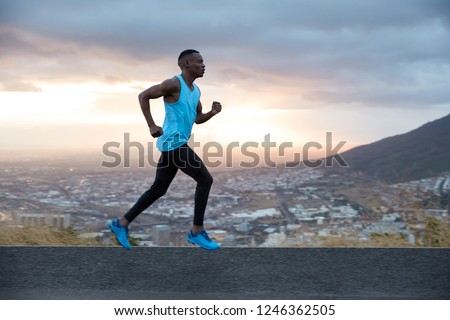 Full length horizontal shot of fast male runner enjoys speed, photographed in motion, runs along road with mountain and city view in background. Running at dawn concept. Strong sportsman outside