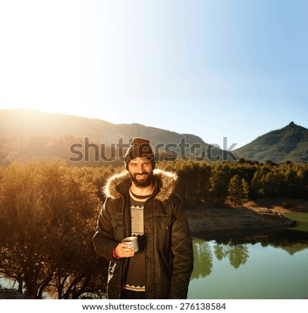 Strong hiker standing on the mountain near lake in background and resting with hot coffee. Happy multiracial smiling man outdoors. Lifestyle photo of Caucasian breaded man.