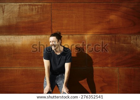 Hipster urban man portrait. Casual dressed man in black t-shirt and washed jeans sly smiling at camera in front of cool textured wooden wall in sunny beautiful day.