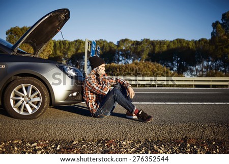 Young brutal man sits near car talking on cell phone because his auto broken down. Caucasian male on a beautiful bright sunny summer day trying to call friend that they help repair car.