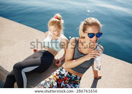 Runner woman drinking water on beach with asian friend running in background. Happy smiling fitness sport model take a break after outdoor workout.