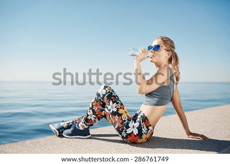 Young fitness blonde woman drinking water after running at beach. Woman sport runner resting taking a break with water bottle drink outside after training.