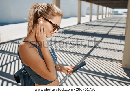 Young fitness blonde woman in sportswear listening music with headphones after training outdoors at beautiful sunny day. Girl runner enjoying the sun and listen music in earphones from smartphone.