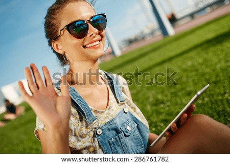 Close up portrait of young blonde woman with tablet. Student girl meets friends outdoor sitting on grass and smiling.