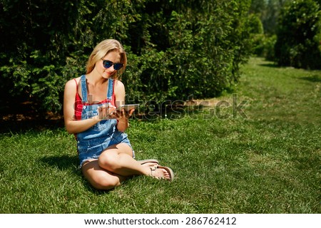 Happy young blonde woman college student use tablet pc sit on grass in park.