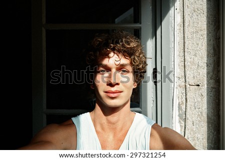 Top view of handsome young curly man making selfie and smiling while standing against window background at sunset time.