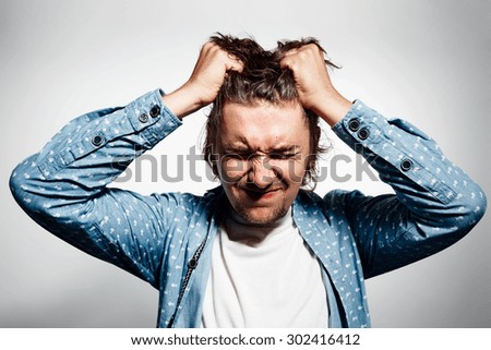 Closeup headshot very sad depressed, stressed disappointed gloomy young man head on hands screaming in despair isolated on grey wall background. Human emotion facial expression reaction