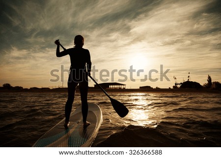silhouette of young girl paddleboarding at sunset, recreation sport paddling ocean beach surf orange sunlight reflection hue on water