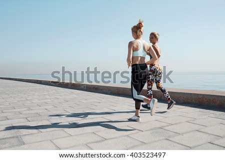 Two female runners exercising outdoors training together for marathon. Caucasian running partners working out outdoors on sunny day. Two fit joggers talking and smiling during workout on the beach.