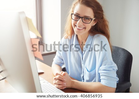 Portrait of happy successful Caucasian businesswoman in blue shirt and glasses looking and smiling at the camera. Attractive female manager sitting at desk after hard working day