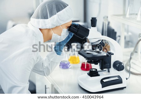 Chemical laboratory scene: young PhD student scientist girl looking through a microscope. Side view of female medical worker in research laboratory (biochemistry, genetics, forensics, microbiology)