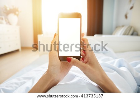 Cropped shot of female holding cell phone with blank copy space screen for your promotional content. Woman using smart phone while lying in bed against home interior background. Film effect, flare sun