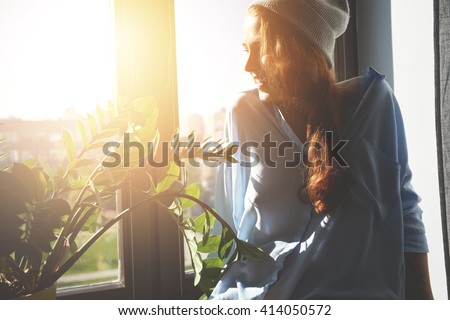 Portrait of happy Caucasian female student spending summer holidays in a big city. Hipster teenager with red hair in gray cap and shirt looking and smiling through the window. Flare sun, film effect