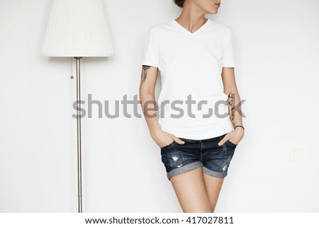 Cropped portrait of attractive young woman wearing blank copy space T-shirt for your text or advertising content, looking away, hands in pockets. Hipster model with tattoos posing isolated indoor
