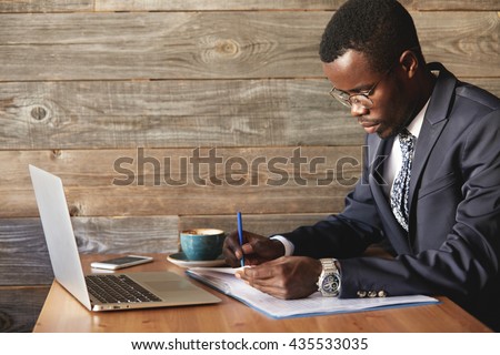 Wealthy and serious dark-skinned businessman with laptop checking report in cafe with a cup of coffee. Young African entrepreneur is focused on work issues and signing papers for business deals.