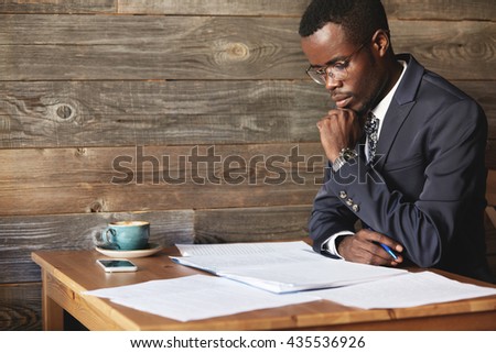 Handsome young African entrepreneur reading a contract before signing it, resting his elbow on the wooden table at a restaurant, sitting with confident concentrated look against wooden wall background