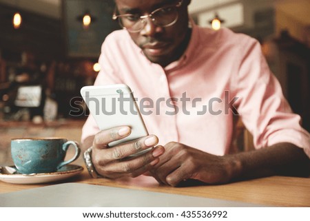 Film effect. Handsome African student in shirt and glasses using mobile phone, looking at the screen with serious and concentrated expression while having coffee at a cafe after classes at university