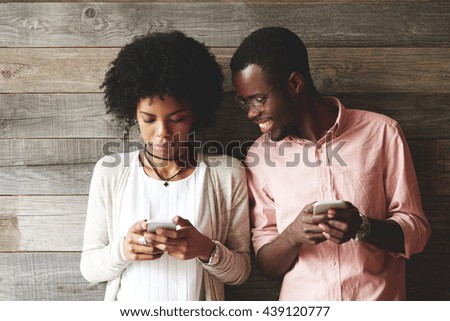 African couple using gadgets: pretty girl with Afro hairstyle in trendy clothes, typing a message on cell phone while her boyfriend standing next to her, looking down at the screen of her smart phone