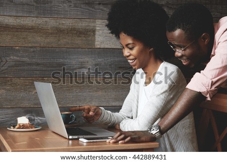 African couple having coffee at a cafe: black girl sitting in front of laptop pointing at the screen and laughing, looking through wedding photos together with her husband who is standing next to her