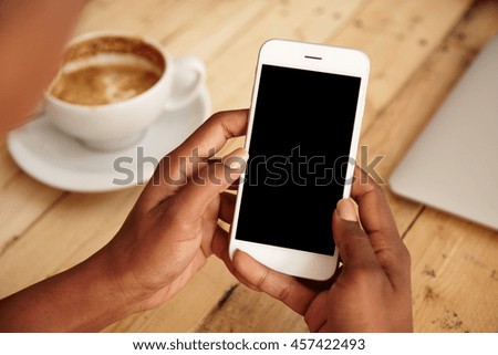 Cropped shot of black female\'s hands holding mobile phone with blank copy space screen for your text message or promotional content. Young woman watching videos using cell phone sitting at cafeteria
