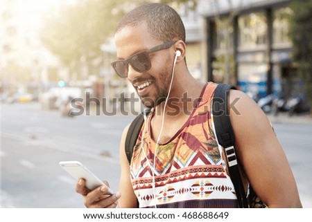 Street fashion. Smiling handsome dark-skinned hipster guy listening to audiobook using his headphones and mobile phone, messaging via social networks, using wireless Internet connection during walk