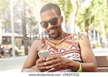 Handsome dark-skinned guy in stylish sunglasses with sly smile flirting online with his girlfriend using web-enabled mobile phone, sitting on bench in city park on hot summer morning, dressed casually