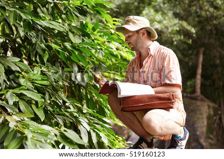 Science and ecology. Handsome bearded ecologist with briefcase and manual exploring problems of wildlife at field work in rainforest, holding leaves of exotic plant, looking serious and concerned