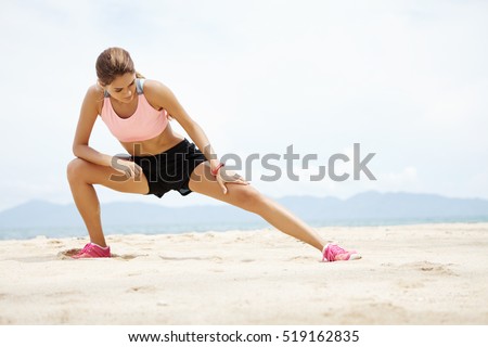 Young female runner with beautiful fit body warming-up her muscles before strength training cardio workout. Woman athlete in sportswear stretching legs with lunge hamstring stretch exercise on beach