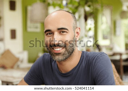 Close up shot of handsome Caucasian man with beard dressed in t-shirt looking and smiling at camera with happy cheerful expression, sitting at sidewalk restaurant on sunny day, waiting for friends