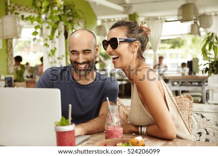 Adorable couple laughing out loud while sitting at outdoor cafe with their modern laptop and watching movies online, using free wi-fi. Happy man and woman looking at computer screen and smiling
