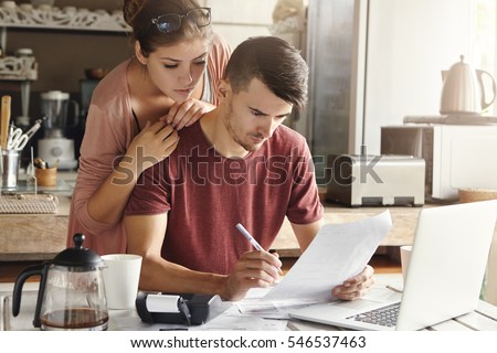 Young unhappy family paying utility bills online on laptop. Stressed man sitting at table with documents and calculator, filling in papers, calculating domestic expenses together with his pretty wife