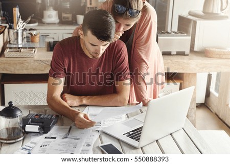 Young family managing budget, reviewing their bank accounts using generic laptop pc and calculator in kitchen. Husband and wife doing paperwork together, paying taxes online on notebook computer