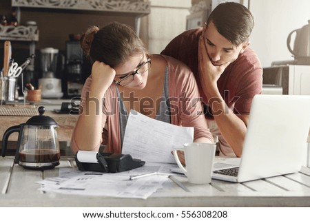 Young stressed Caucasian couple facing financials troubles, sitting at kitchen table with papers, calculator and laptop computer and reading document from bank, looking frustrated and unhappy