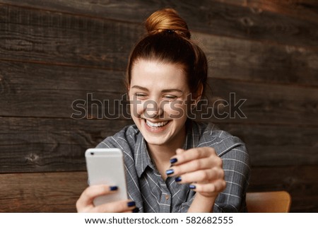 Cheerful young redhead female student with cute smile siting in modern cafe interior, using cell phone, checking newsfeed on her social network accounts. Pretty girl surfing internet on mobile