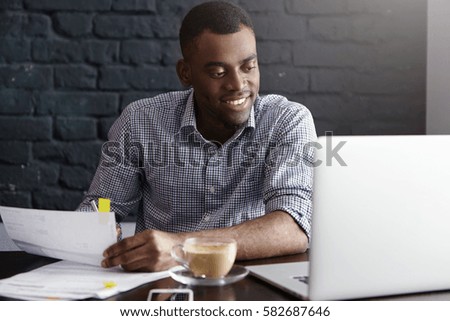 Happy confident young African-American businessman in formal wear filling in papers while managing finances at cafe during lunch break, sitting at table in front of open laptop, having cappuccino