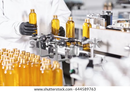 Automated process of production new cosmetic products on big modern factory. Scientist standing near conveyor line putting glass bottles with yellow thick substance on it. Production of new shampoo