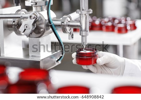 Production and packaging of cosmetics products. Cropped shot of factory worker in gloves and gown holding plastic jar, filling it with white face or body cream. Beauty, skincare and cosmetology