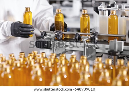 Plastic bottles with yellow liquid substance of shampoo, shower gel or soap standing in rows on assembly line at factory. Researcher in black gloves working at production line in modern laboratory