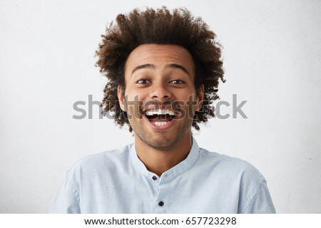 Carefree joyful handsome Afro American man with bushy hairstyle and bristle having shining eyes opening his mouth with joy bursting into laughing. Positive human expressions, emotions and feelings