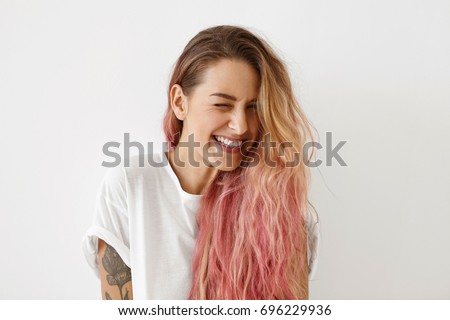 Young female model with pink hair tips, winking her eyes while having joy, smiling broadly, feeling joy. Beautiful hipster female with long hair feeling happiness while being photographed in studio