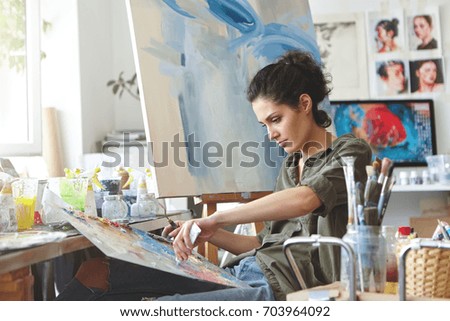 Young female student having classes at art studio, learning how to draw landscapes, trying to mix different watercolors on cardboard. Concentrated woman with dark hair, dressed casually, painting